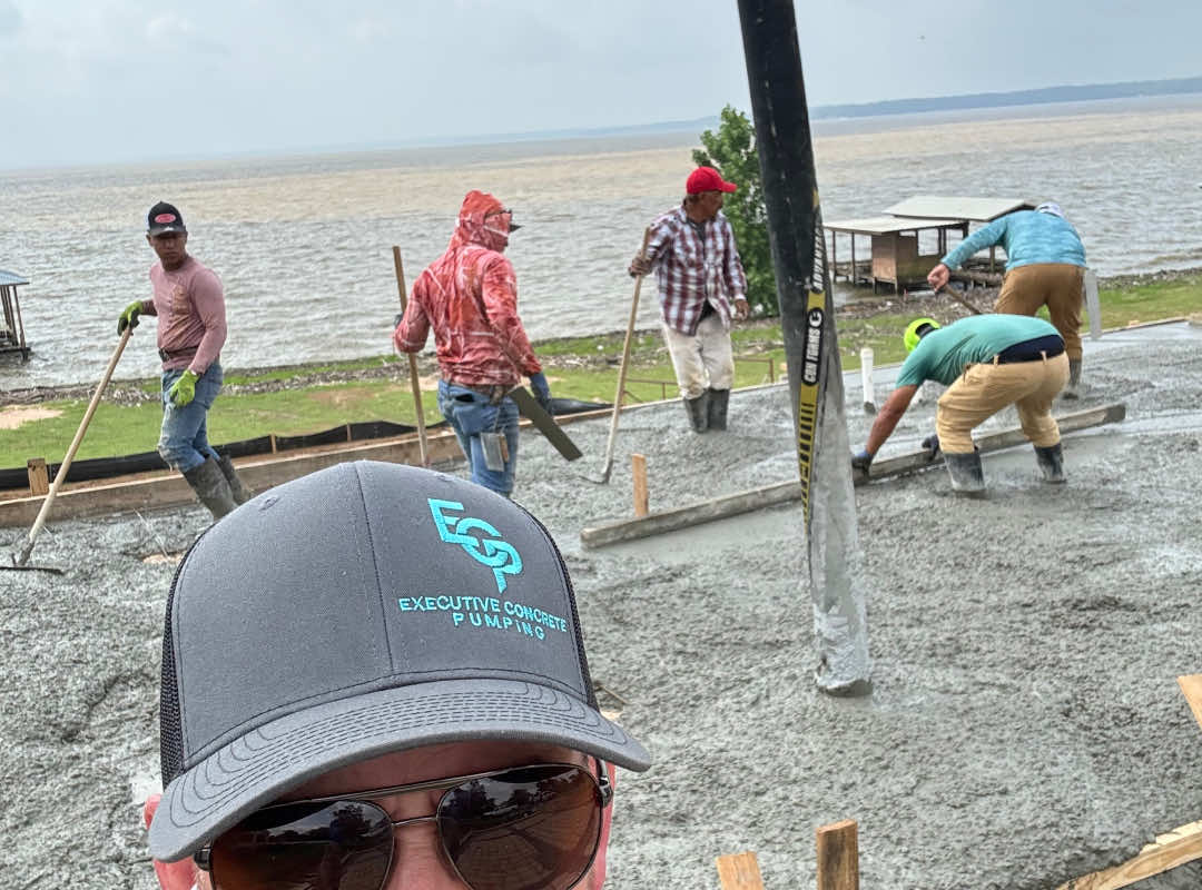 Team of workers leveling freshly poured concrete near a lakeside, with a visible Executive Concrete Pumping logo on a cap in the foreground.