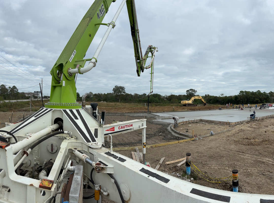 Close-up of a green boom pump pouring concrete at a construction site, with workers leveling a freshly poured slab in the background under a cloudy sky.