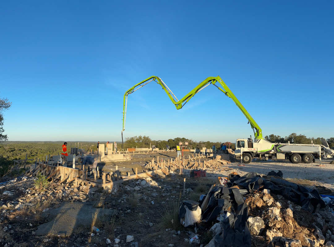 Nighttime construction site with a green boom pump illuminated by work lights, as workers pour and level concrete on a prepared foundation.