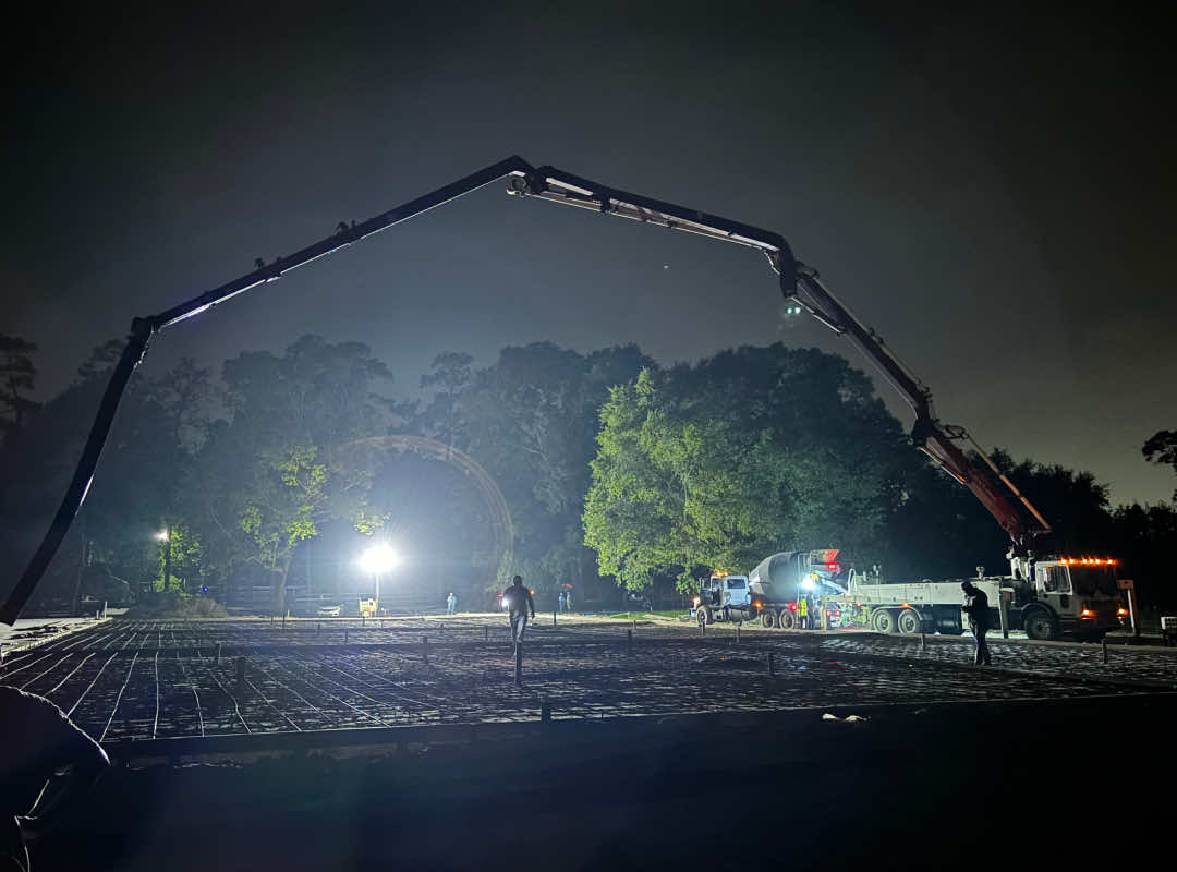 Nighttime concrete pour at a construction site with illuminated equipment, including a large boom pump and cement trucks, under bright work lights and surrounded by trees.