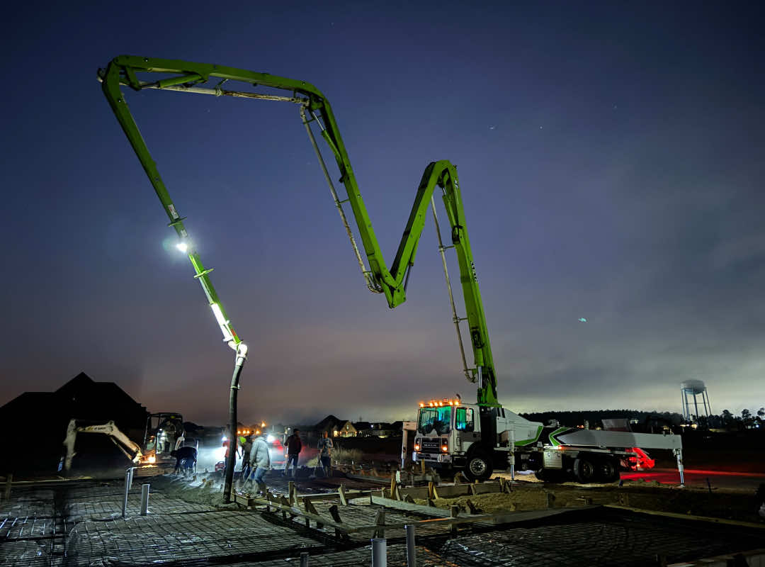 Nighttime construction site with a green boom pump illuminated by work lights, as workers pour and level concrete on a prepared foundation.