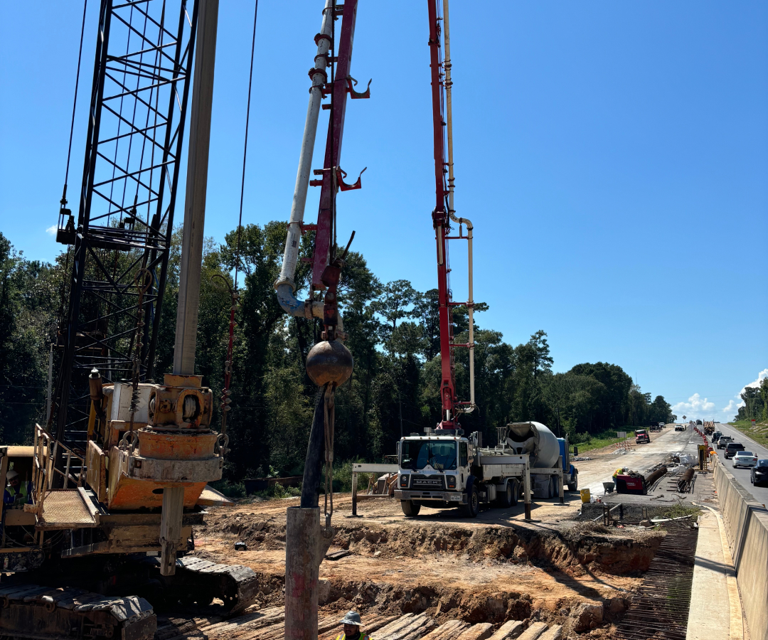 Construction site by a highway with a red concrete pump and heavy machinery working on foundation pouring under a clear blue sky, Houston TX Concrete Pumping and Delivery Services