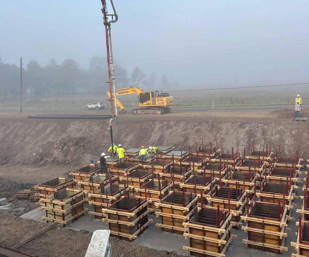 Construction site on a foggy morning featuring a yellow excavator and workers filling foundation molds with a red concrete pump, Houston TX Concrete Pumping and Delivery Services