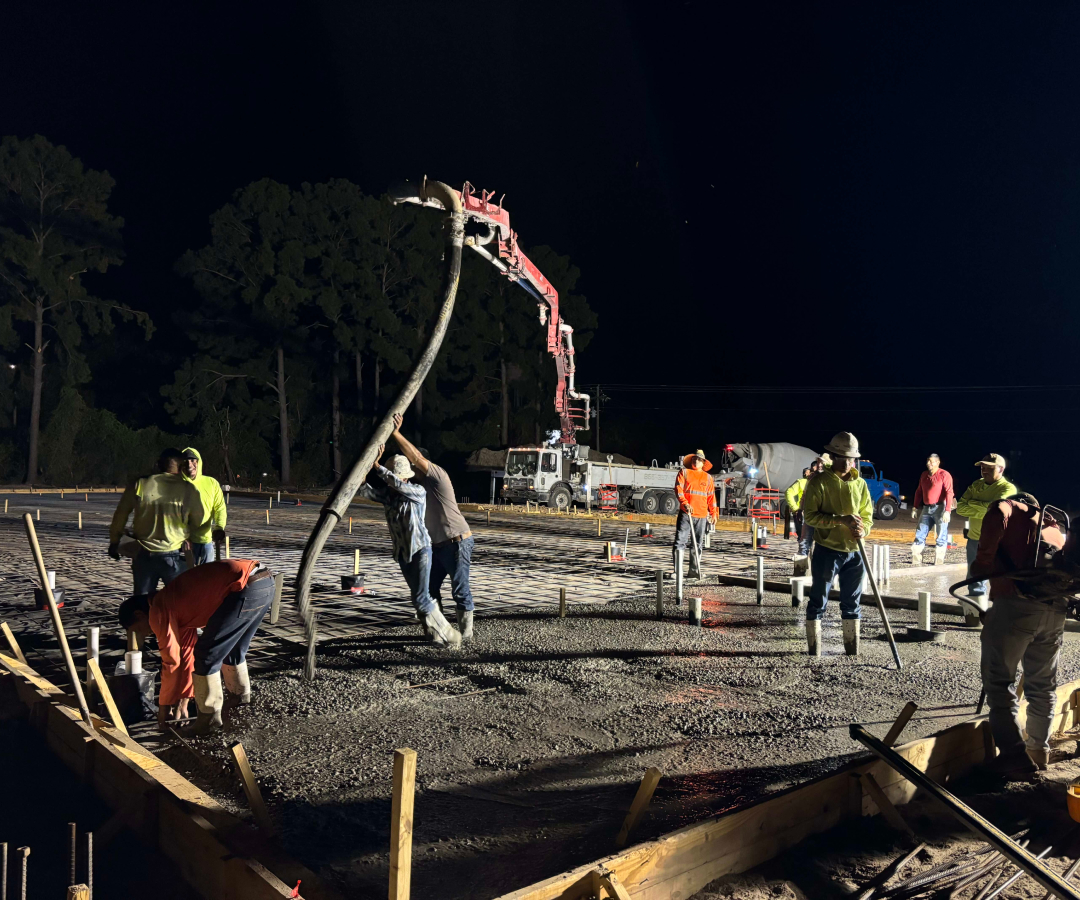 Nighttime concrete pour with workers leveling the slab and a red boom pump illuminated under work lights at the construction site, Houston TX Concrete Pumping and Delivery Services