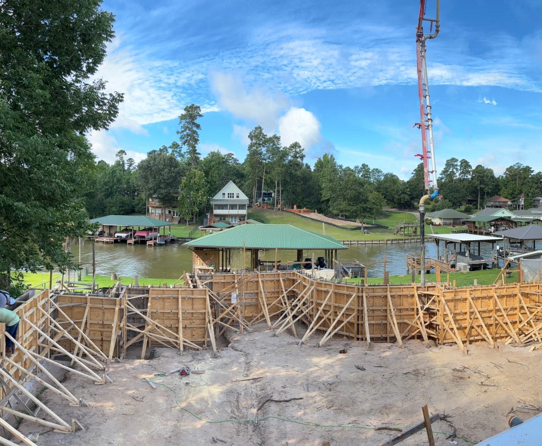 Concrete pour in progress at a lakeside property construction site, surrounded by wooden forms and a view of lakefront homes, Houston TX Concrete Pumping and Delivery Services